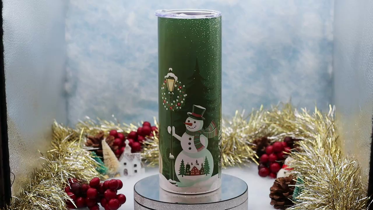Video Showcasing A hand holds a green insulated tumbler decorated with a winter scene featuring a snowman, Christmas wreath, street lamp, evergreen trees, and small houses