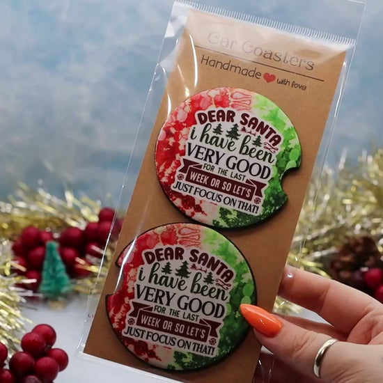 video showcasing a two round car coasters with a holiday-themed design. The coasters feature a watercolor Christmas tree and the text 'DEAR SANTA i have been VERY GOOD FOR THE LAST WEEK ON SO LET'S JUST FOCUS ON THAT!'.