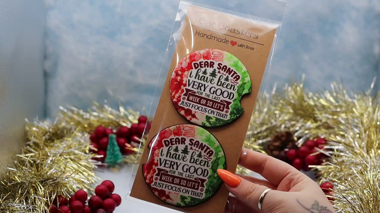 video showcasing a two round car coasters with a holiday-themed design. The coasters feature a watercolor Christmas tree and the text 'DEAR SANTA i have been VERY GOOD FOR THE LAST WEEK ON SO LET'S JUST FOCUS ON THAT!'.