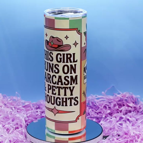Video showcasing A hand holds a tall insulated tumbler with a printed western-themed design. The tumbler has a cream background with brown text reading 'THIS GIRL RUNS ON SARCASM & PETTY THOUGHTS,