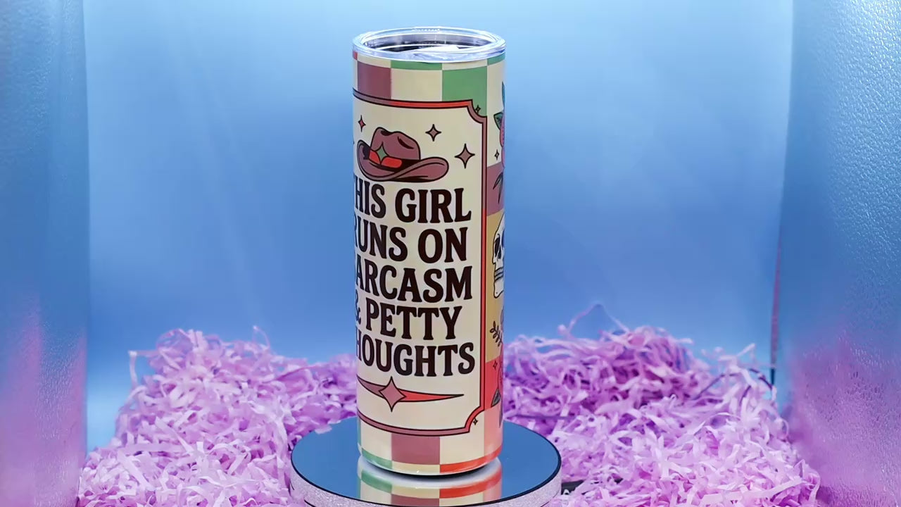 Video showcasing A hand holds a tall insulated tumbler with a printed western-themed design. The tumbler has a cream background with brown text reading 'THIS GIRL RUNS ON SARCASM & PETTY THOUGHTS,