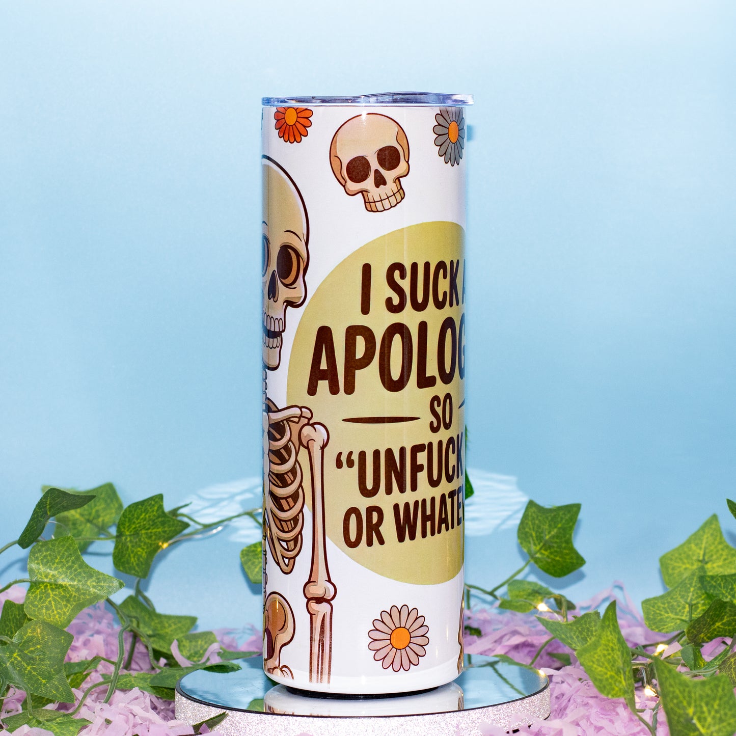 A white insulated tumbler with a humorous skeleton graphic and text reading 'I SUCK APOLOGIES SO "UNFUCK YOURSELF OR WHATEVER"', displayed on a small mirrored pedestal with green ivy and purple flowers against a light blue background.