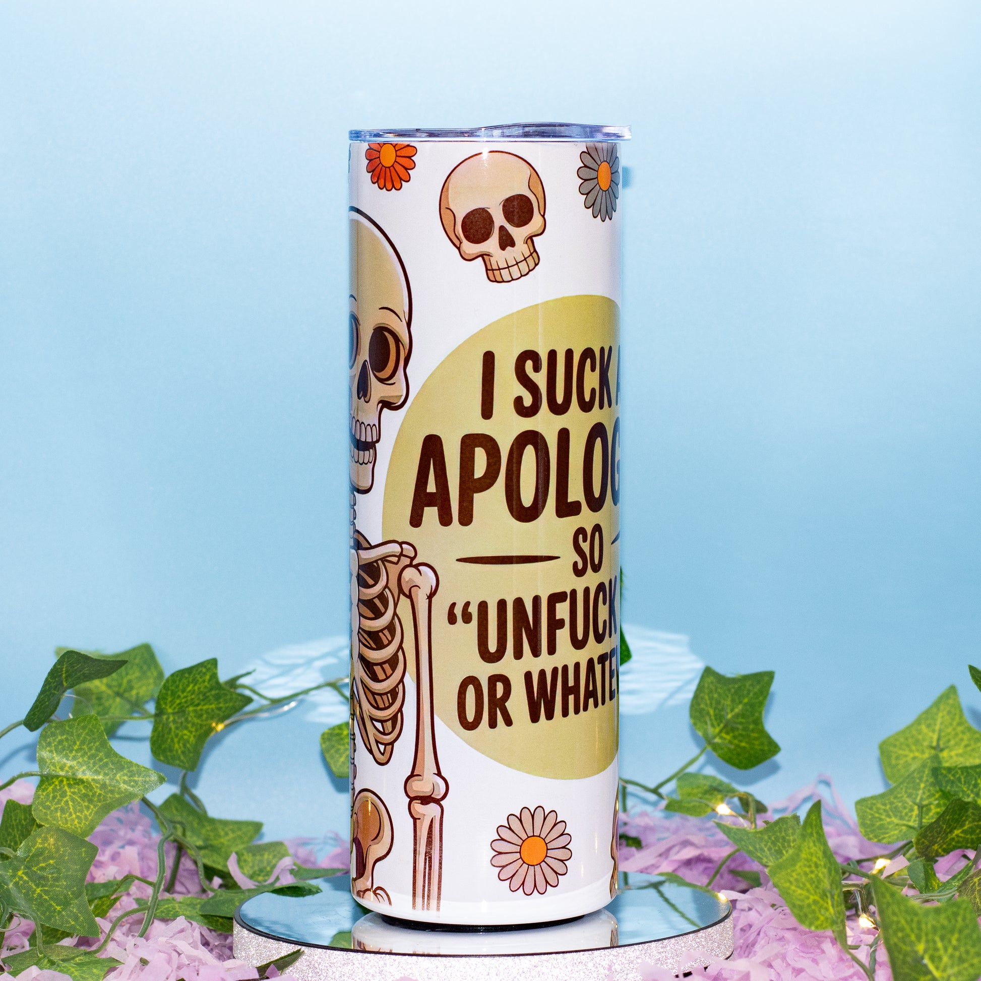 A white insulated tumbler with a humorous skeleton graphic and text reading 'I SUCK APOLOGIES SO "UNFUCK YOURSELF OR WHATEVER"', displayed on a small mirrored pedestal with green ivy and purple flowers against a light blue background.