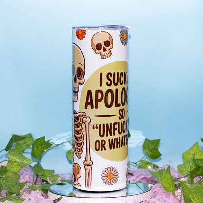 A white insulated tumbler with a humorous skeleton graphic and text reading 'I SUCK APOLOGIES SO "UNFUCK YOURSELF OR WHATEVER"', displayed on a small mirrored pedestal with green ivy and purple flowers against a light blue background.