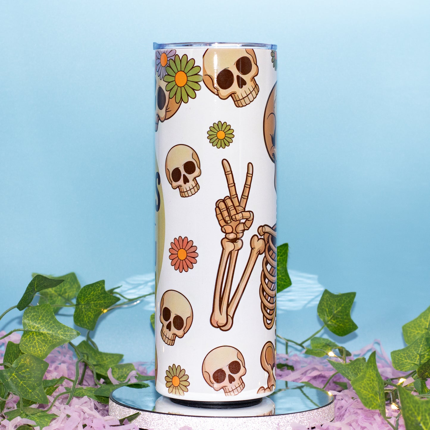 A white insulated tumbler with a Day of the Dead style print featuring skulls, bones, and flowers, displayed on a mirrored surface with green ivy and purple petals against a light blue background.