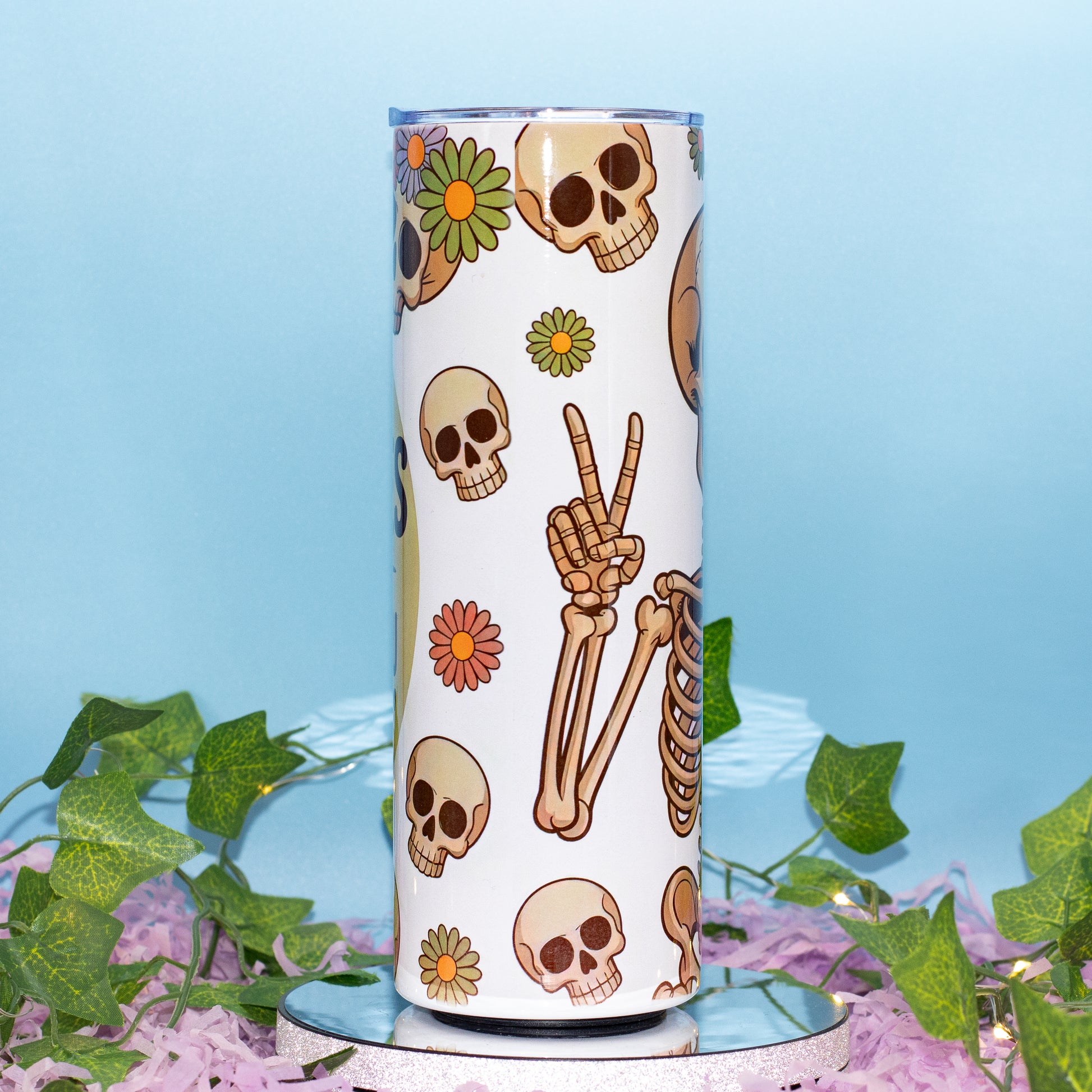 A white insulated tumbler with a Day of the Dead style print featuring skulls, bones, and flowers, displayed on a mirrored surface with green ivy and purple petals against a light blue background.