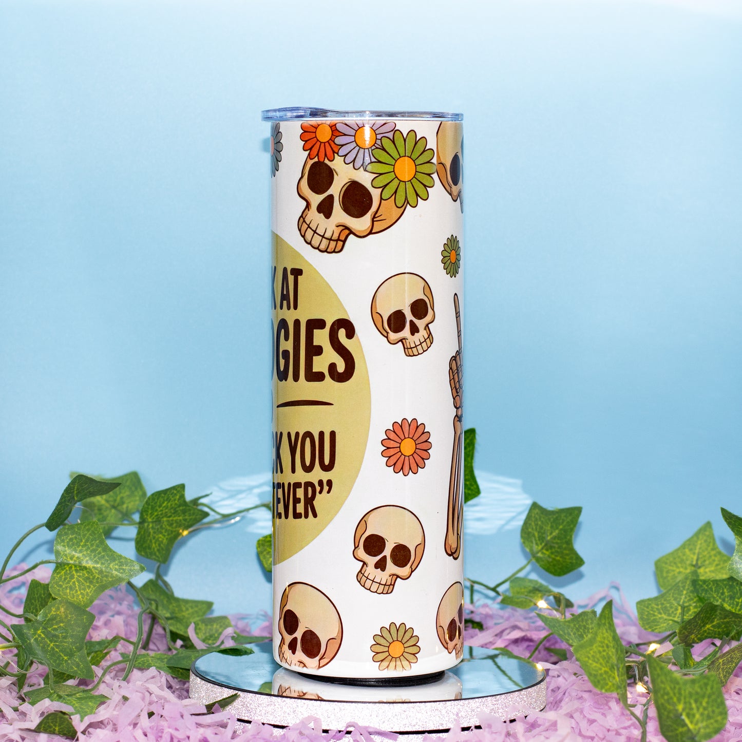 A white insulated tumbler with a Day of the Dead theme, featuring sugar skull illustrations, colorful flowers, The tumbler is displayed on a small mirrored stand surrounded by artificial green ivy and pink petals against a light blue background.