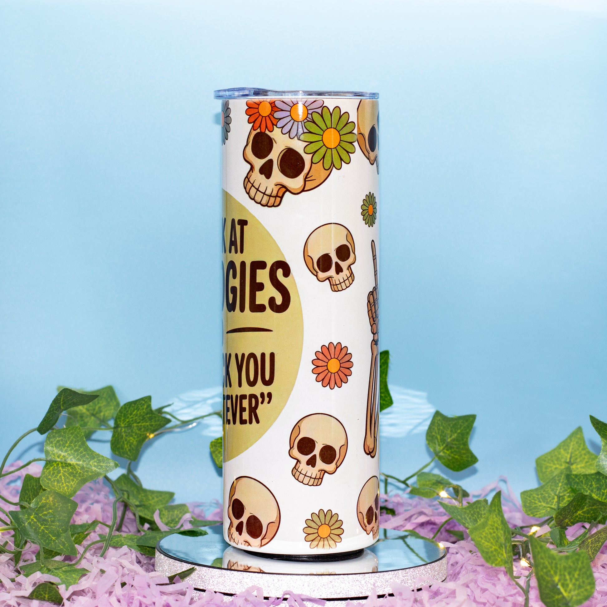 A white insulated tumbler with a Day of the Dead theme, featuring sugar skull illustrations, colorful flowers, The tumbler is displayed on a small mirrored stand surrounded by artificial green ivy and pink petals against a light blue background.
