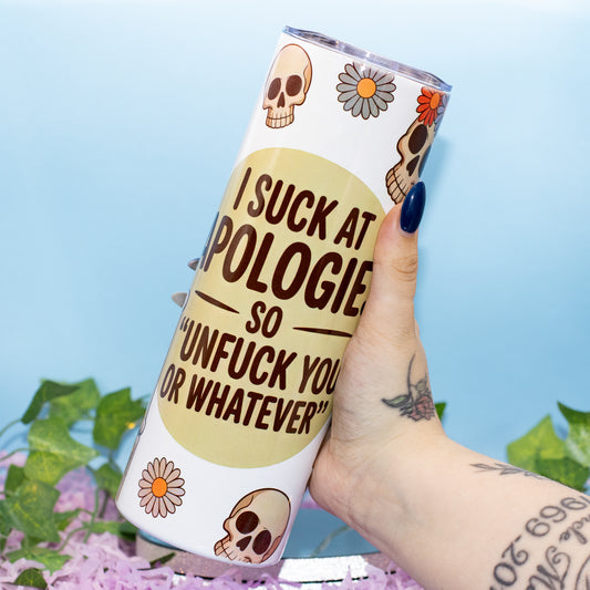 A hand holds a white insulated tumbler with a humorous apology-themed slogan, decorated with skull and daisy illustrations, against a light blue background with decorative green leaves and purple flowers.