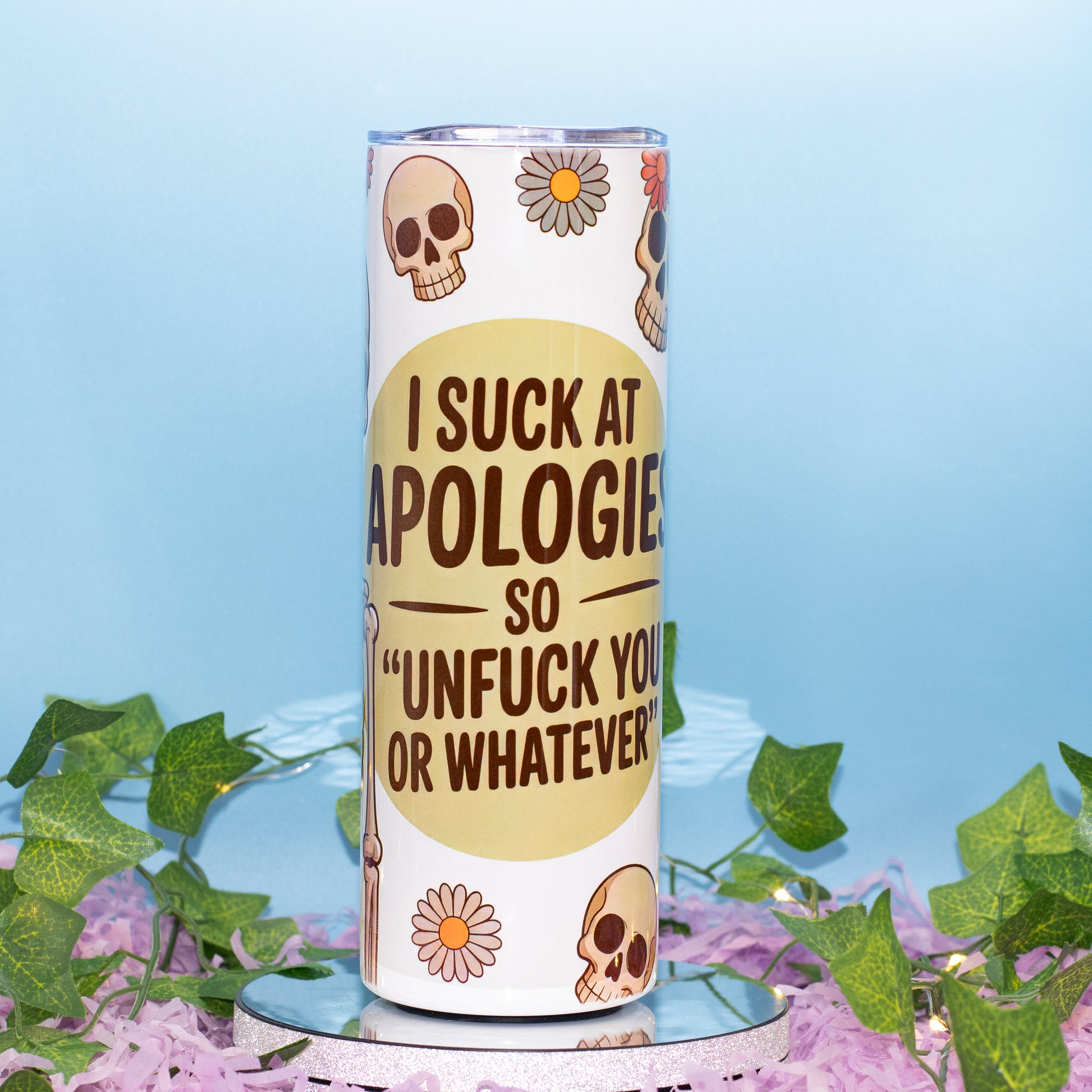 A white insulated tumbler with a humorous message printed on it, styled with decorative green ivy and purple flowers on a light blue background.