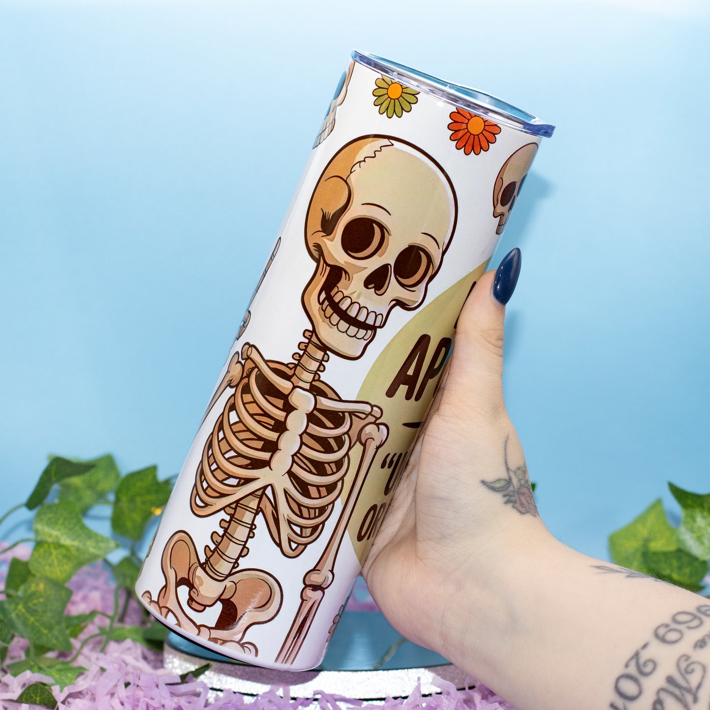A hand holds a white insulated tumbler featuring a cartoon skeleton design and floral accents against a light blue background with decorative green leaves and purple flowers.