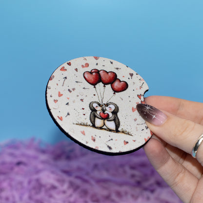 A hand holds a round white coaster featuring a cute cartoon penguin holding red heart-shaped balloons. The background is a bright blue sky with purple fluffy paper at the bottom.