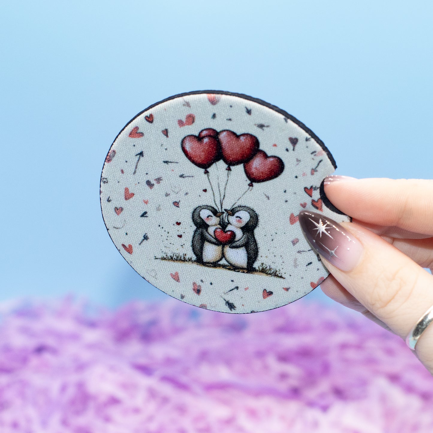 A hand holding a round car coaster featuring a cute illustration of two penguins holding red heart-shaped balloons, with scattered small hearts on a light background, set against a soft blue and purple blurred backdrop.