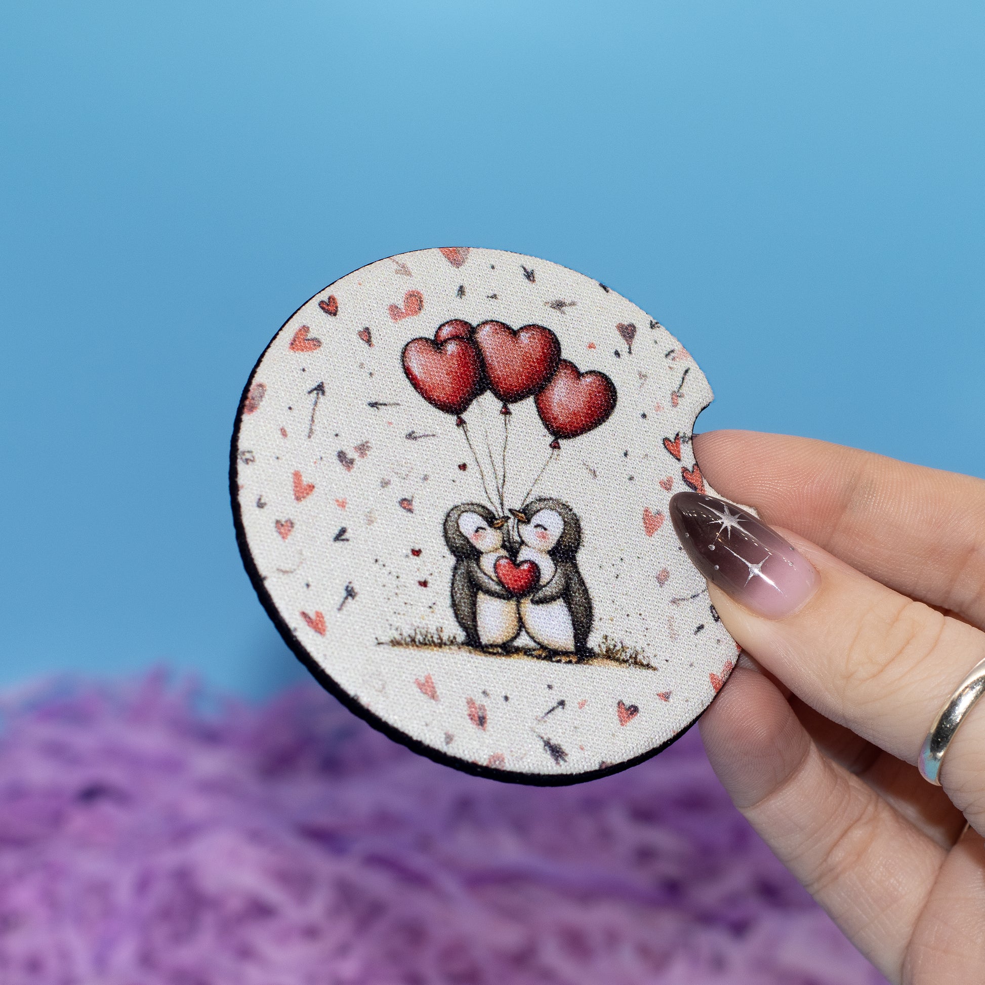 A hand holds a round car coaster featuring a cute illustration of two penguins holding red heart-shaped balloons. The coaster has a speckled off-white background with small red hearts