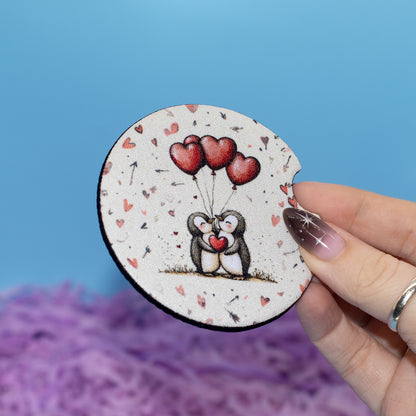 A hand holds a round car coaster featuring a cute illustration of two penguins holding red heart-shaped balloons. The coaster has a speckled off-white background with small red hearts