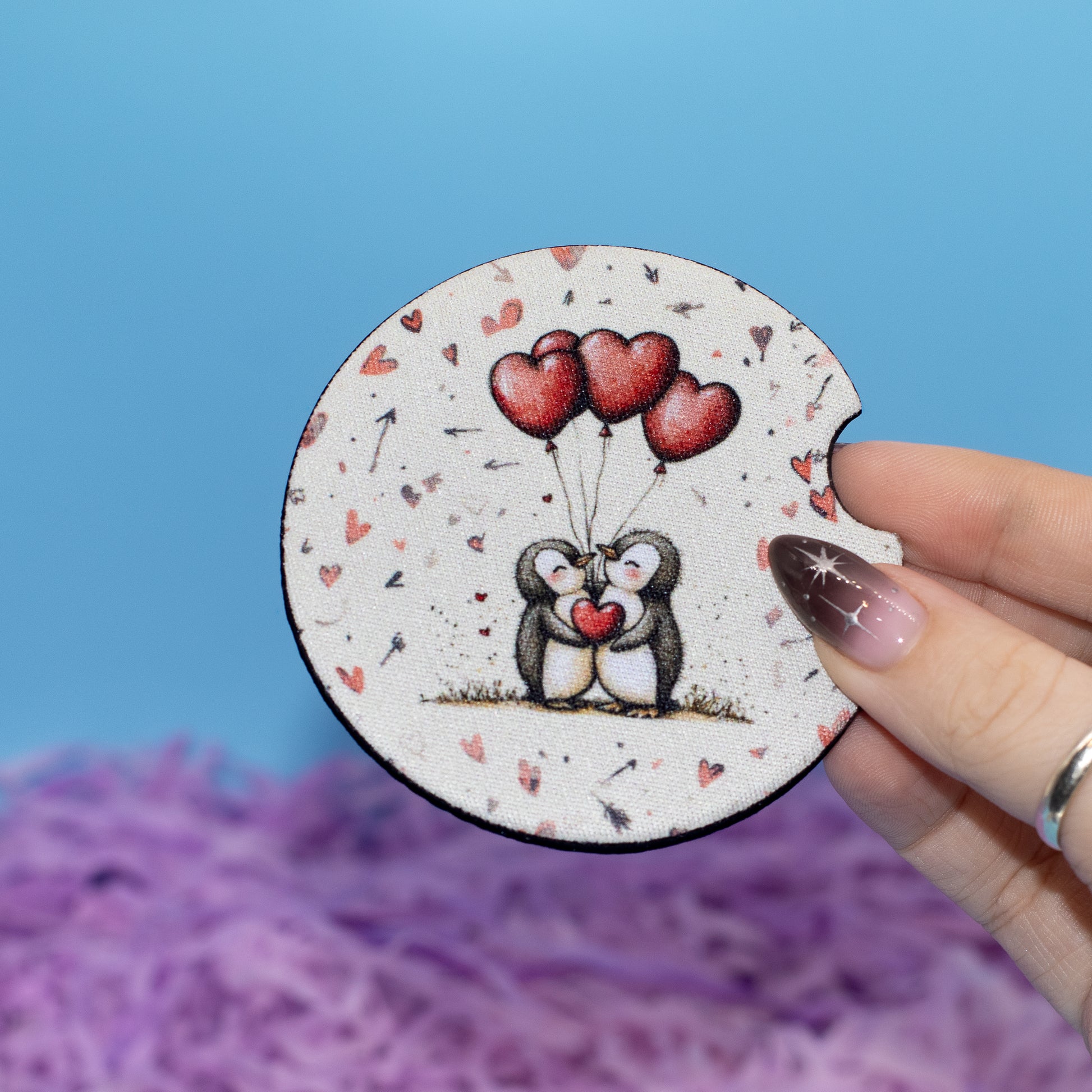 A hand holds a round coaster featuring a cute illustration of two penguins holding red heart-shaped balloons. The coaster has a white background with small red hearts and black speckles, set against a blue background with purple shredded paper.