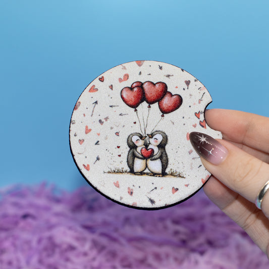 A hand holds a round coaster featuring a cute illustration of two penguins holding red heart-shaped balloons. The coaster has a white background with small red hearts and black speckles, set against a blue background with purple shredded paper.