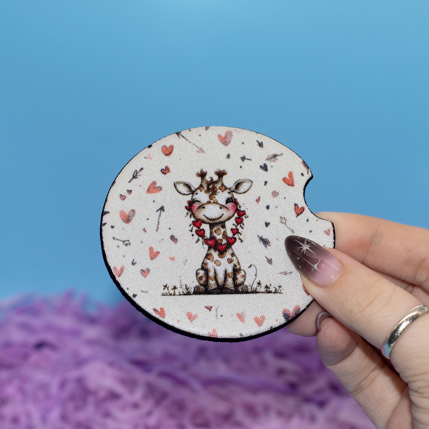 A hand holds a round neoprene coaster featuring a cute giraffe illustration with a red floral collar and hearts, set against a blue background with purple shredded paper.