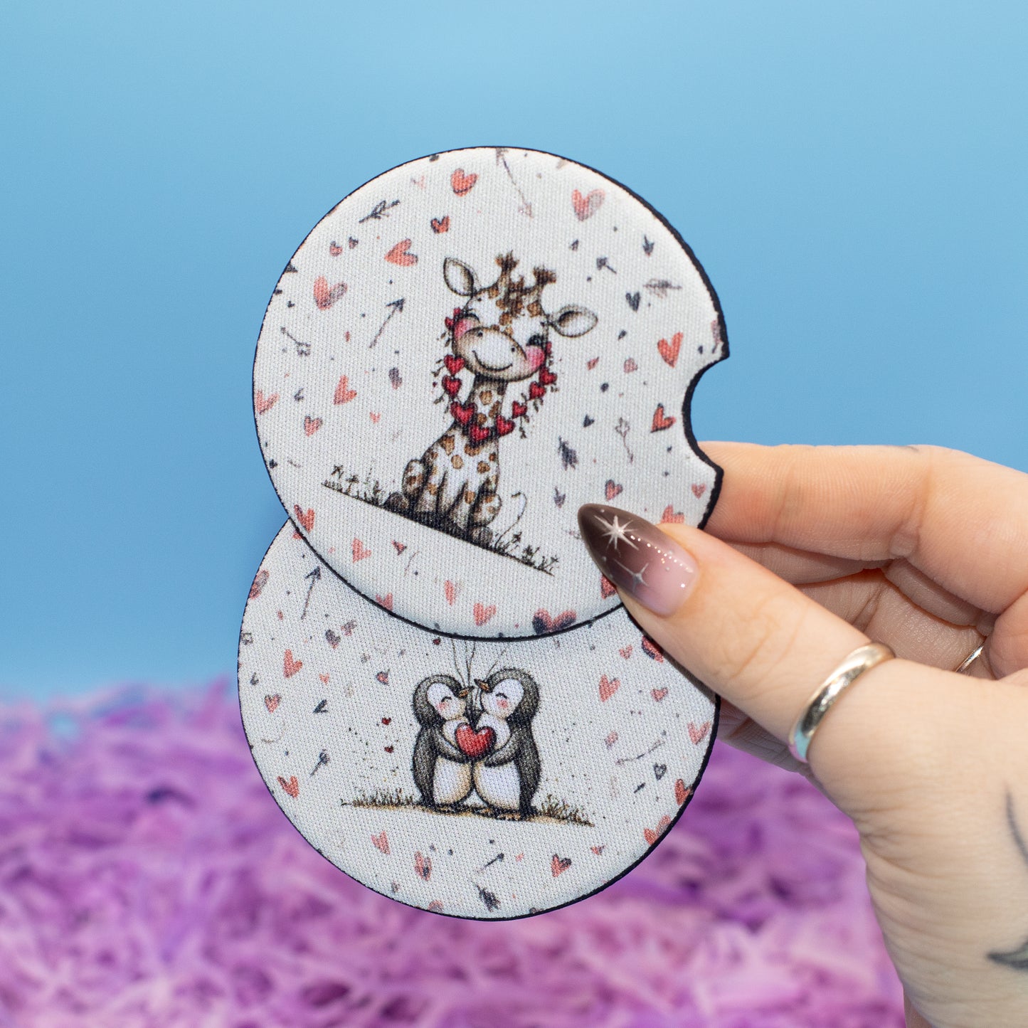 Two round car coasters with cute animal illustrations (a giraffe and a penguin) on a white background with pink hearts, held by a hand against a blue and purple background.